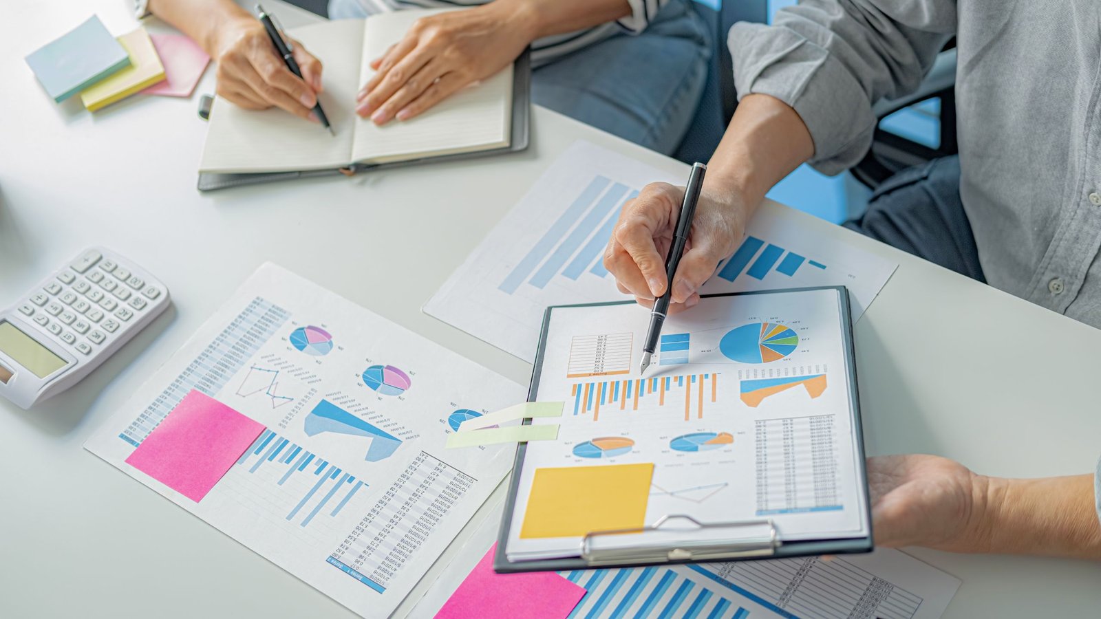 image showing Two professionals analyzing startup growth charts and business data with graphs, pie charts, and a calculator on a white desk.